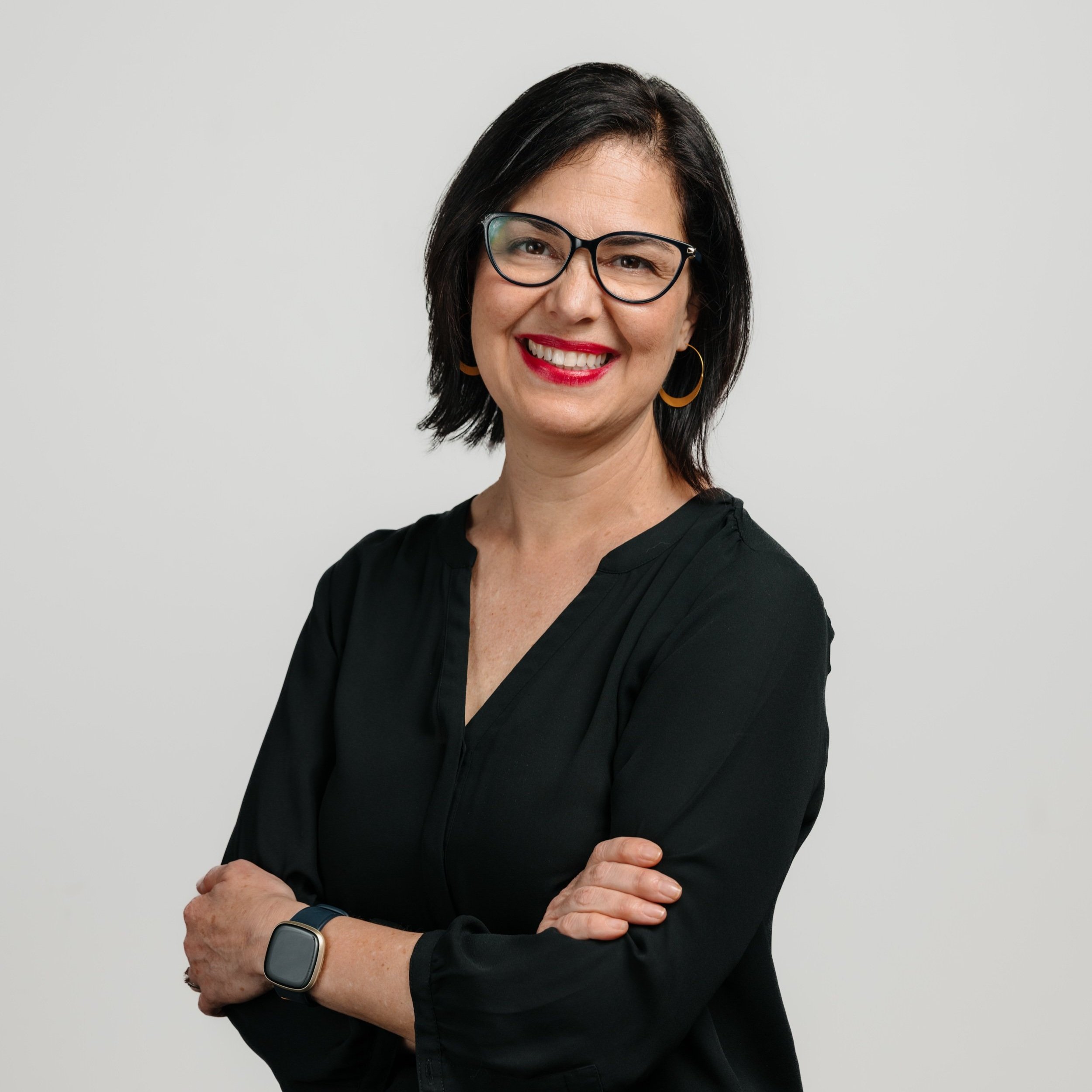 A smiling portrait of Tammy, who has black shoulder-length hair and brown eyes. She is Latina wearing red lipstick, gold hoops, black cat-eye glasses, a black quarter-sleeve v-neck top, and an Apple watch.