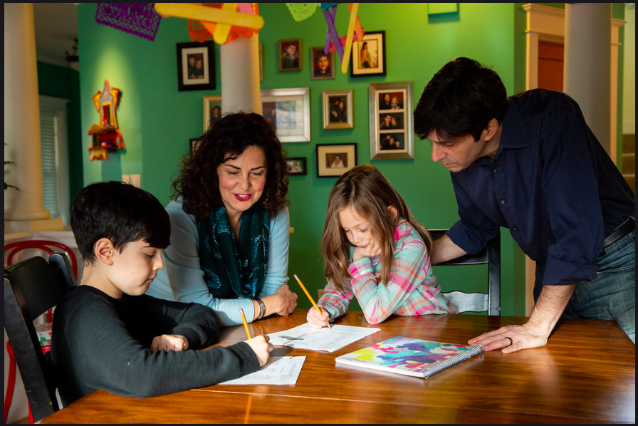 A photo of Tammy and her husband leaning over a dining room table while two of their younger kids work on homework
